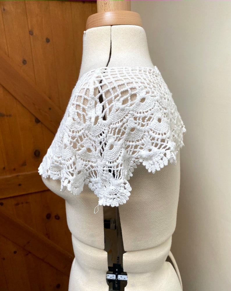May include: White crochet lace collar with a delicate floral pattern. The collar is displayed on a white dress form.