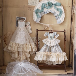 Puede incluir: Una exhibici&oacute;n de ropa de mu&ntilde;eca vintage, que incluye dos vestidos, un gorro y un velo. Los vestidos presentan intrincados detalles de encaje y volantes, uno en blanco y otro en azul claro. El gorro tiene una cinta azul y el velo es de tela transparente.