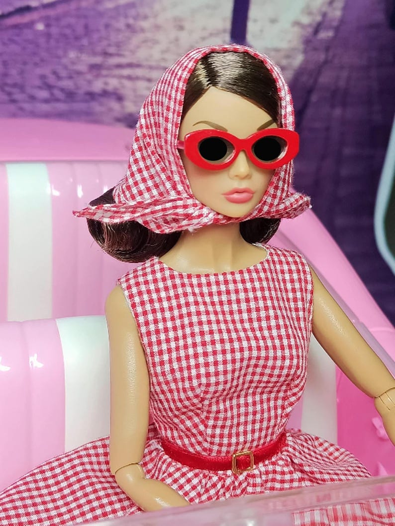 K&ouml;nnte beinhalten: Eine Puppe tr&auml;gt ein rot-wei&szlig; kariertes Kleid mit einem roten G&uuml;rtel und einem rot-wei&szlig; karierten Kopftuch. Die Puppe tr&auml;gt rote ovale Sonnenbrillen und hat braunes Haar.
