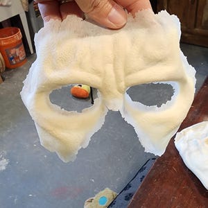 Peut inclure: Un masque cr&egrave;me avec des ouvertures pour les yeux, tenu. Le masque a une surface textur&eacute;e et est probablement fait d'un mat&eacute;riau souple. Le masque est probablement destin&eacute; &agrave; un costume ou &agrave; un usage th&eacute;&acirc;tral.