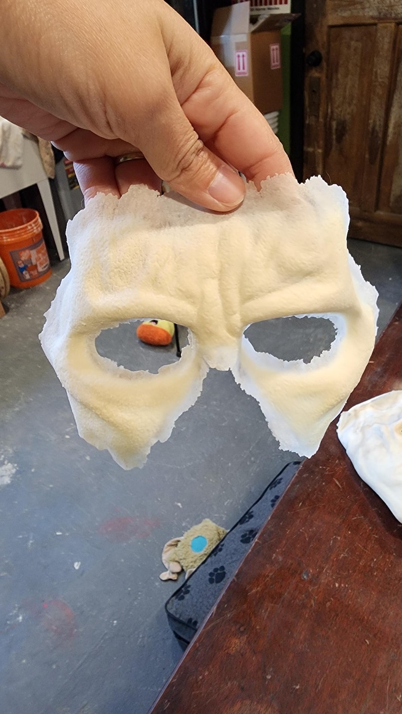 Peut inclure: Un masque cr&egrave;me avec des ouvertures pour les yeux, tenu. Le masque a une surface textur&eacute;e et est probablement fait d'un mat&eacute;riau souple. Le masque est probablement destin&eacute; &agrave; un costume ou &agrave; un usage th&eacute;&acirc;tral.
