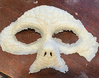 Peut inclure: Un masque crème avec un motif texturé ressemblant à celui d'un reptile. Le masque comporte des ouvertures pour les yeux, un nez et une surface détaillée. Le masque est probablement destiné à un costume ou à un usage théâtral.