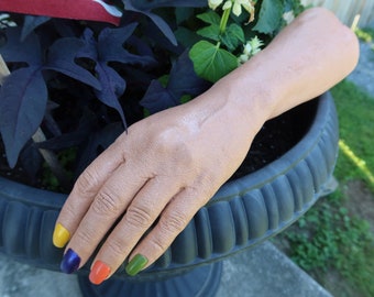 Peut inclure: Un modèle de bras et de main prothétique réaliste grandeur nature avec un teint de peau clair. Les doigts présentent des ongles peints de couleurs distinctes : jaune, violet foncé, orange et vert olive. La main repose sur le bord d'un pot.