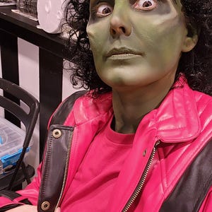 Peut inclure: Une personne avec du maquillage vert sur le visage et des cheveux boucl&eacute;s fonc&eacute;s, portant une veste rouge avec des accents noirs. La veste a une fermeture &eacute;clair et une chemise rouge en dessous. La personne regarde la cam&eacute;ra.