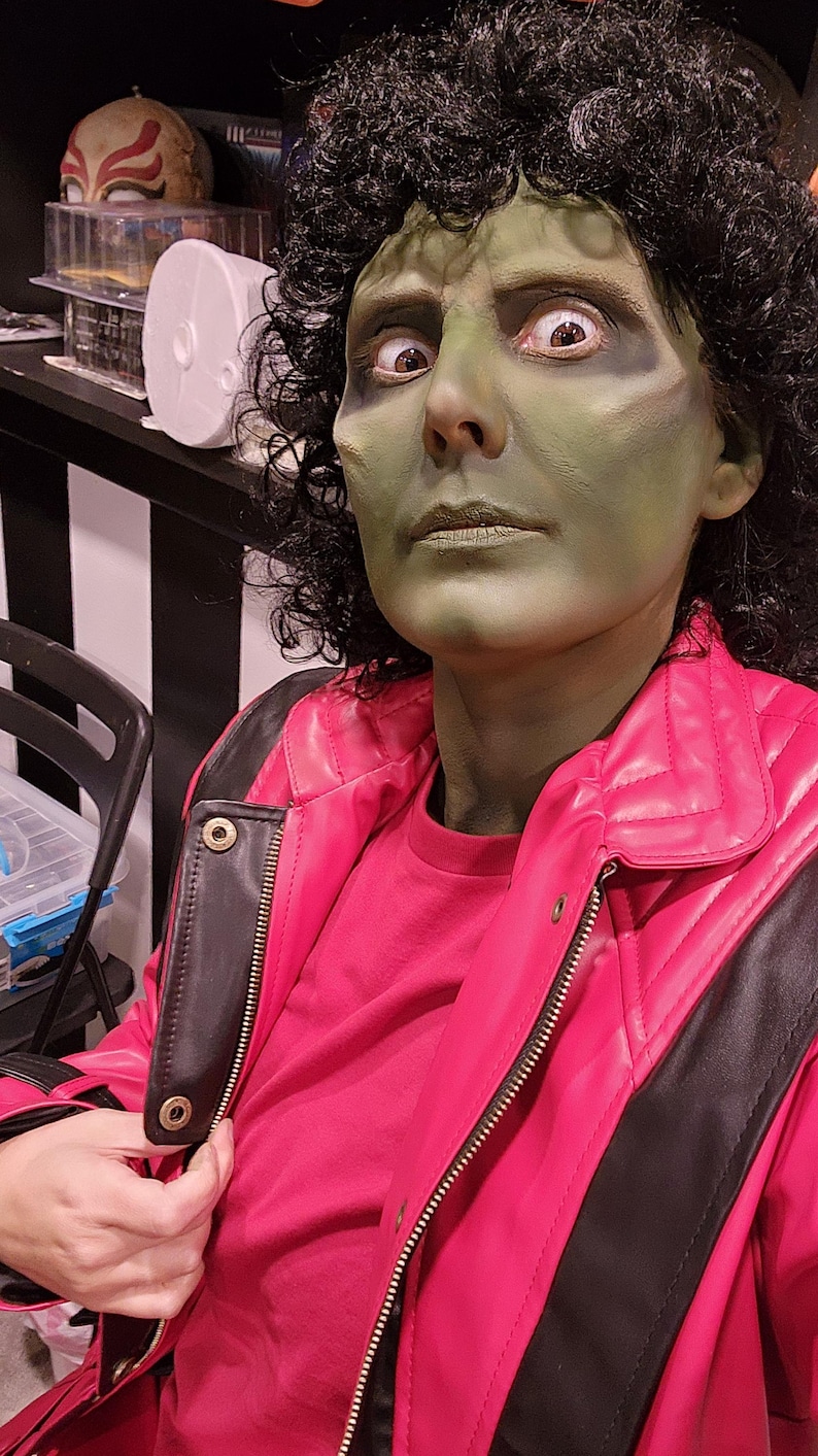 Peut inclure: Une personne avec du maquillage vert sur le visage et des cheveux boucl&eacute;s fonc&eacute;s, portant une veste rouge avec des accents noirs. La veste a une fermeture &eacute;clair et une chemise rouge en dessous. La personne regarde la cam&eacute;ra.