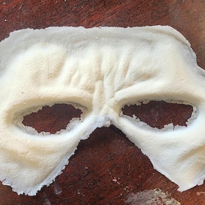 Peut inclure: Un masque cr&egrave;me avec des ouvertures pour les yeux, probablement pour un costume ou un usage th&eacute;&acirc;tral. Le masque a une surface textur&eacute;e et repose sur une surface en bois sombre. La forme du masque sugg&egrave;re qu'il couvre le haut du visage.