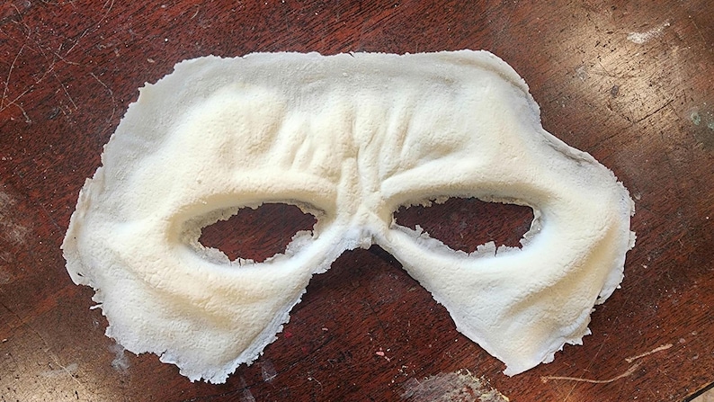 Peut inclure: Un masque cr&egrave;me avec des ouvertures pour les yeux, probablement pour un costume ou un usage th&eacute;&acirc;tral. Le masque a une surface textur&eacute;e et repose sur une surface en bois sombre. La forme du masque sugg&egrave;re qu'il couvre le haut du visage.