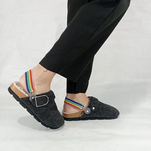 May include: Gray felt clogs with a rainbow strap and a cork sole. The clogs have a buckle closure and a round toe.