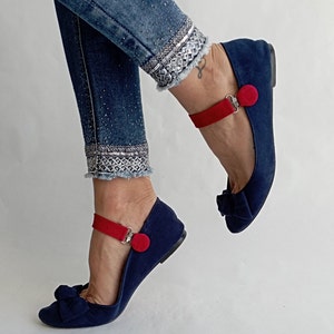 Könnte beinhalten: Ein Paar marineblaue Ballerinas mit roten Riemen und einem roten Knopf als Akzent. Die Ballerinas haben eine Schleife an der Spitze.