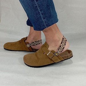 Peut inclure: Sabots en daim marron avec une boucle de fermeture et un lit de pied en liège. Les sabots ont une sangle de cheville à motif géométrique.