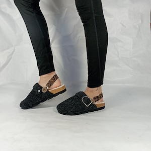 May include: Dark charcoal gray felt clogs featuring a round toe, a cork-style midsole, and a black rubber outsole. The footwear includes a leopard print heel strap with a metallic buckle and a soft, light-colored faux fur lining for comfort.