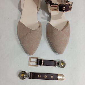 May include: A pair of beige flats with a pointed toe and a brown ankle strap with a gold buckle. Two brown belts with gold buckles and decorative accents are also displayed.