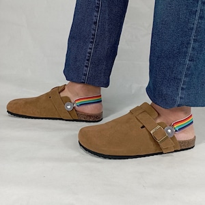 Könnte beinhalten: Braune Wildledersandalen mit Schnallenverschluss und einem regenbogenfarbenen Gummiband. Die Sandalen haben ein Korkfußbett und eine schwarze Laufsohle.
