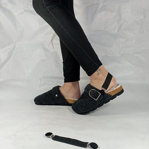 May include: A pair of gray fleece lined clogs with a black adjustable strap and buckle. The clogs have a cork footbed and a black elastic strap with metal clips.