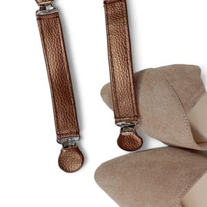 May include: Two pairs of copper-colored leather shoe clips with silver metal clasps. The clips are attached to a pair of beige suede shoes.