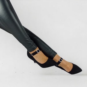May include: A pair of black ankle strap heels with a pointed toe. The heels are worn by a person wearing black leather pants.