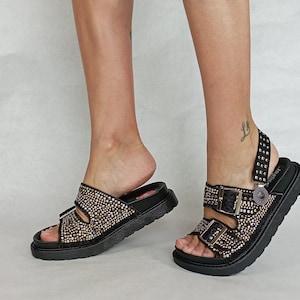 May include: Black platform sandals with two adjustable buckles and a rhinestone-covered strap. The sandals have a chunky platform sole and a back strap with a decorative metal charm.