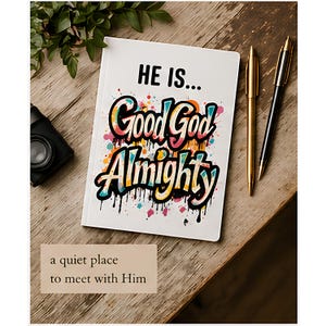 May include: A white journal with the words "HE IS... Good God Almighty" in colourful graffiti-style lettering. Two pens, one gold and one black, lie beside the journal on a wooden surface. A small, black camera is also visible.