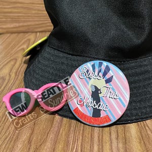 May include: A black bucket hat, pink sunglasses, and a round button with the text "Bless This Mosaic" and "Hot Rat Summer" are displayed. The sunglasses have a rectangular frame. The button has a pink, blue, and white striped background.