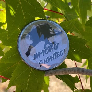 May include: A circular pinback button featuring a black silhouette of a creature running against a light blue background. The text JIMOTHY SIGHTED! is printed in bold black capital letters across the lower portion of the circular surface.
