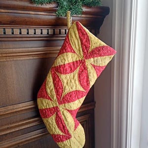 May include: A quilted Christmas stocking in red and yellow. The stocking features a repeating pattern of red petal-like shapes on a yellow background. The stocking is hanging on a wooden surface, with a green garland above.
