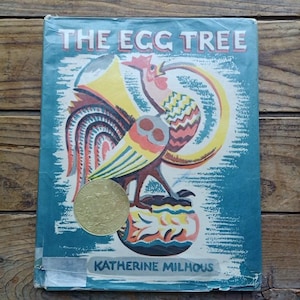 May include: A vintage book cover for "The Egg Tree" by Katherine Milhous. The cover displays a colourful illustration of a rooster, owl, and decorative elements in yellow, red, and blue. A gold coin is placed on the cover.