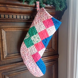 May include: A patchwork Christmas stocking in shades of pink, green, red, and blue. The stocking is made of quilted fabric and hangs from a wooden cabinet. The top and hanging loop are made of a pink floral fabric. The stocking is approximately 40 cm long.
