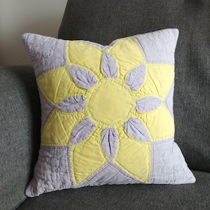 May include: A square pillow with a yellow and purple quilted design. The design features a large yellow flower with purple petals.