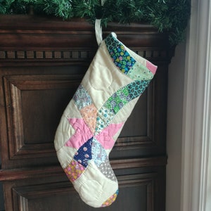May include: A white quilted Christmas stocking with a patchwork design featuring pink, blue, green, and brown fabric. The stocking is hanging on a dark wood surface.