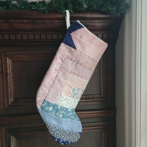 Peut inclure: Une chaussette de Noël faite à la main avec un motif patchwork. Elle présente divers tissus pastel, notamment roses et bleus, avec des motifs floraux. Le haut est bleu marine et une boucle blanche permet de l'accrocher. Fond en bois sombre.
