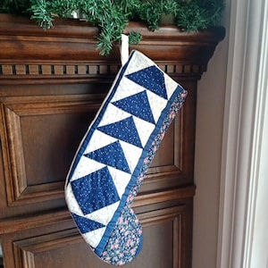 May include: A handmade Christmas stocking with a quilted design. The stocking features a pattern of blue triangles and squares on a white background. The cuff and heel are adorned with a floral print, and a blue ribbon loop is attached for hanging.