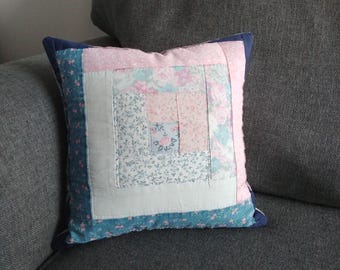 May include: A square patchwork pillow with a log cabin pattern in shades of pink, blue, and white. The pillow has a navy blue border.