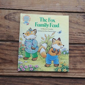 May include: A children's book titled "The Fox Family Feud" by Maureen Connellan, illustrated by Diane Dawson Hearn. The cover features two foxes, one wearing a blue and pink sweater and the other wearing blue overalls and a yellow and orange striped shirt.