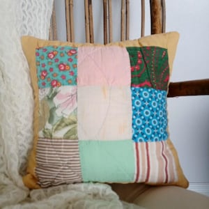 May include: Square patchwork pillow with a yellow border. The pillow features nine fabric squares in various patterns and colors, including floral, striped, and solid pastel hues. The pillow is placed on a wooden chair with a cream-colored lace throw.