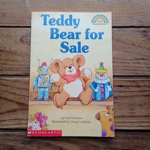 May include: A children's book titled "Teddy Bear for Sale" with illustrations of a teddy bear, robot, and clown. The book cover is yellow with blue text and the Scholastic logo.