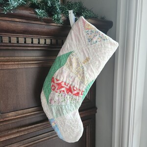 Peut inclure: Une chaussette de Noël matelassée fabriquée à partir de divers morceaux de tissu dans des tons de blanc, vert, rouge, jaune et bleu. La chaussette présente un motif en patchwork avec des imprimés floraux et géométriques, des coutures visibles.