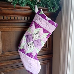 May include: A patchwork Christmas stocking in shades of purple, lavender, and green. The design incorporates geometric patterns and a quilted texture on the toe and cuff. A loop is attached for hanging. The stocking is a festive decoration.