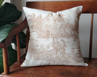 May include: A white throw pillow with a brown toile pattern featuring a farmhouse, trees, and figures. The pillow is sitting on a wooden bench.