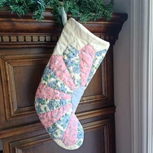 May include: A quilted Christmas stocking featuring a fan-shaped pattern with alternating panels of solid light pink fabric and a floral print fabric in shades of blue, green, and white. The stocking has a cream-coloured cuff and a hanging loop.