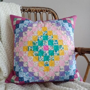May include: Square patchwork pillow with a vibrant, colorful design. The pillow features a grid pattern of small squares in shades of pink, blue, yellow, green, and purple. The pillow is resting on a wicker chair.