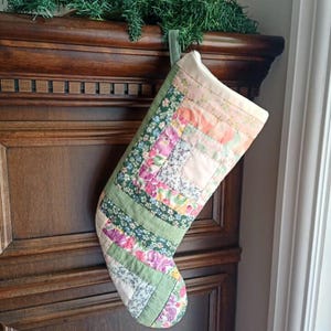 May include: A quilted Christmas stocking constructed from various fabric strips in floral and solid patterns. The patchwork design features shades of pink, green, white, and yellow, with a green fabric loop at the top for hanging purposes.