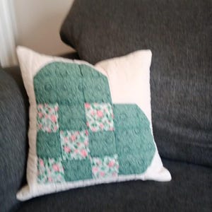 Peut inclure: Un coussin carré avec un motif de cœur en patchwork sur un fond crème. Le cœur est composé d'une alternance de carrés de tissu vert foncé uni et de tissu vert clair orné d'un petit motif floral rose sur toute la surface.