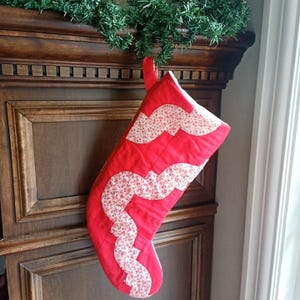 Peut inclure: Une chaussette de Noël matelassée en tissu rouge vif ornée de deux bandes ondulées et festonnées en tissu blanc à petites fleurs rouges. La chaussette possède une boucle de suspension en tissu rouge assorti sur le dessus.
