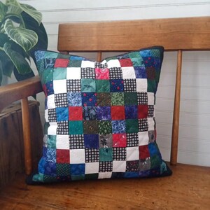 Peut inclure: Coussin patchwork carré avec divers motifs de tissus rouges, verts, bleus et blancs. Le coussin est posé sur une chaise en bois, mettant en valeur sa qualité artisanale et ses couleurs vives. Bordure noire.