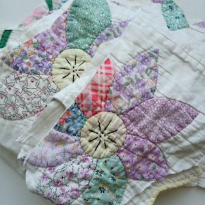 Peut inclure: Un textile matelassé présentant un motif floral avec des pétales et des centres en tissu blanc. Les pétales sont composés de divers tissus à motifs dans des tons de violet, vert, rouge et blanc. Les centres sont jaune pâle avec des détails cousus noirs.