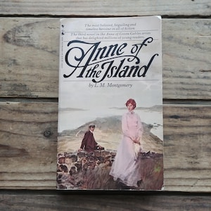 May include: A book cover with a light blue background featuring a woman in a pink dress standing on a stone wall. The title of the book is "Anne of the Island" by L.M. Montgomery.