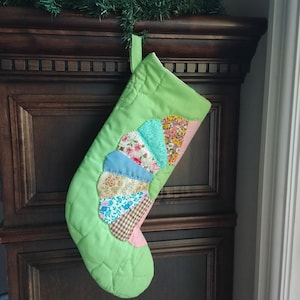 Könnte beinhalten: Ein grüner, gesteppter Weihnachtssocken mit einem Patchwork-Muster aus bunten Stoffresten. Der Socken hängt an einem Holzmantel.
