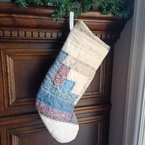 May include: A patchwork Christmas stocking in shades of blue, white, and floral patterns. The quilted stocking hangs from a wooden surface, with a green garland above. The design is reminiscent of a traditional quilt.