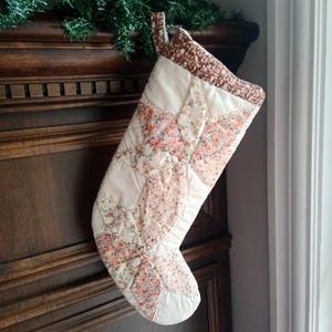 May include: A quilted Christmas stocking featuring a patchwork design of cream-coloured fabric and floral patterned fabric in shades of pink, orange, and green. The stocking has a brown and white floral cuff and a matching fabric hanging loop.