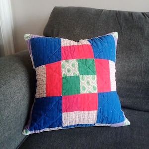 May include: Square patchwork pillow with a quilted design. The pillow features a vibrant pattern of blue, red, green, and white squares. The pillow is placed on a gray sofa.